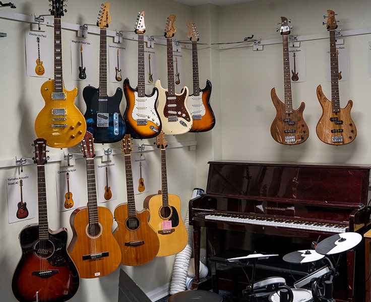 Image of our music classroom including several electric and acoustic guitars hanging on the wall, and an acoustic piano and electric drum kit beside them.
