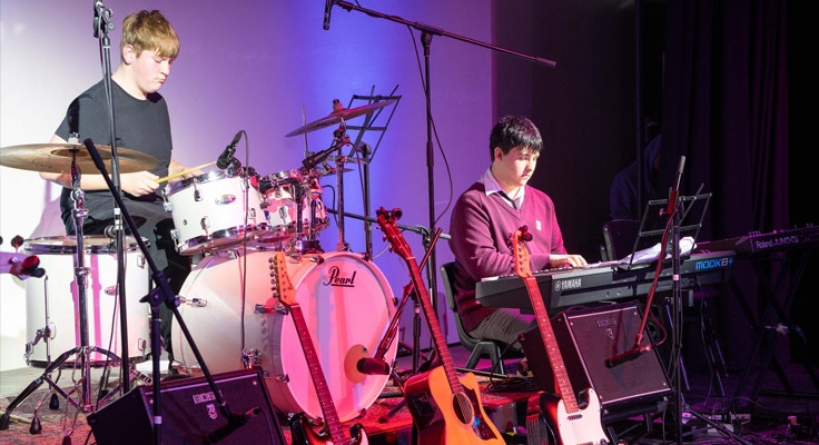 Image of two students performing in a band on stage - one on drum kit and one on keyboard.