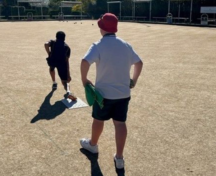 Two students on the green as one of them rolls a lawn bowl.