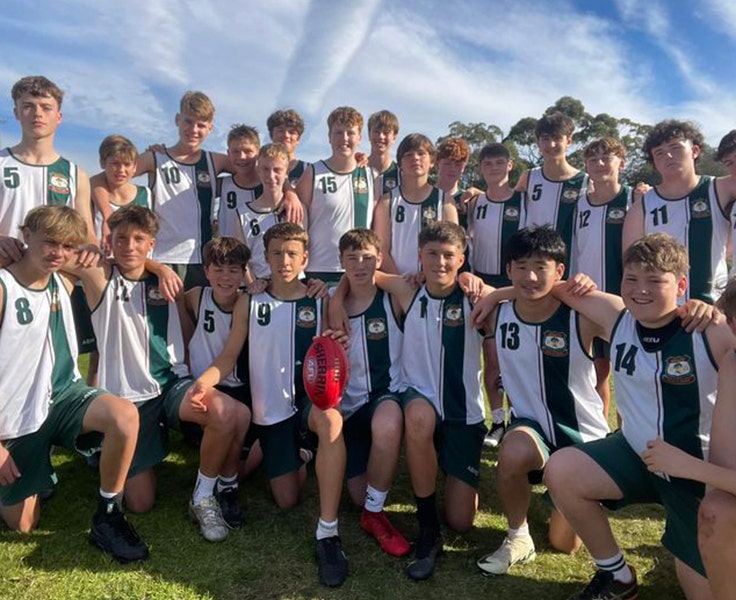 A group of students in our sports uniform jersey/singlet huddled together and smiling with one student front and centre holding an AFL football.