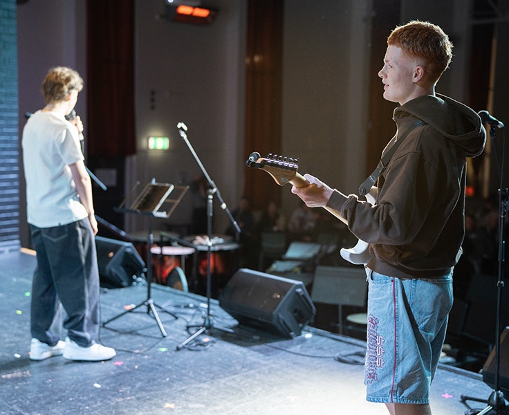 An image of two students - one singer and one electric guitarist on stage performing in a band.