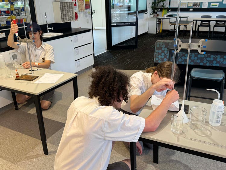 Three students participating in a chemistry experiment involving beakers and retort stands.