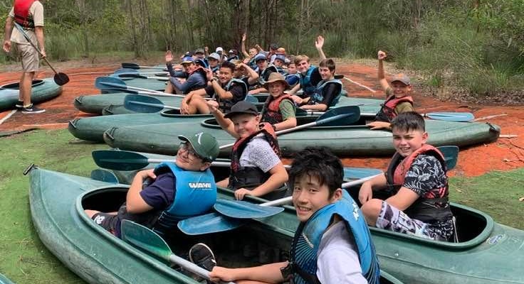 A group of students in kayaks smiling and waving their hands in the air.